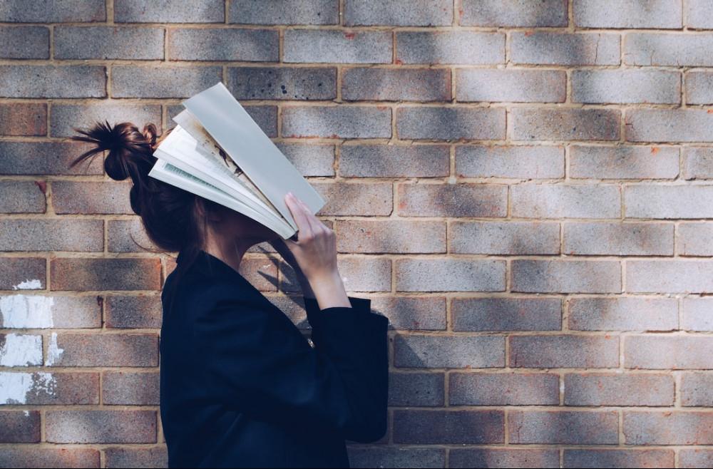 Person frustrated with learning which can be hard Woman with folders over her face standing next to a brick wall