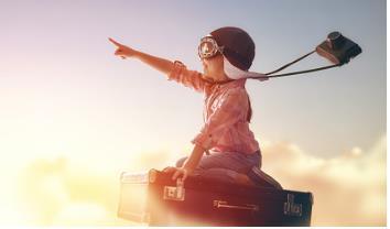 Image success Child with helmet, goggles and camera flying in the clouds sitting on a suitcase while pointing and imaging success in life.