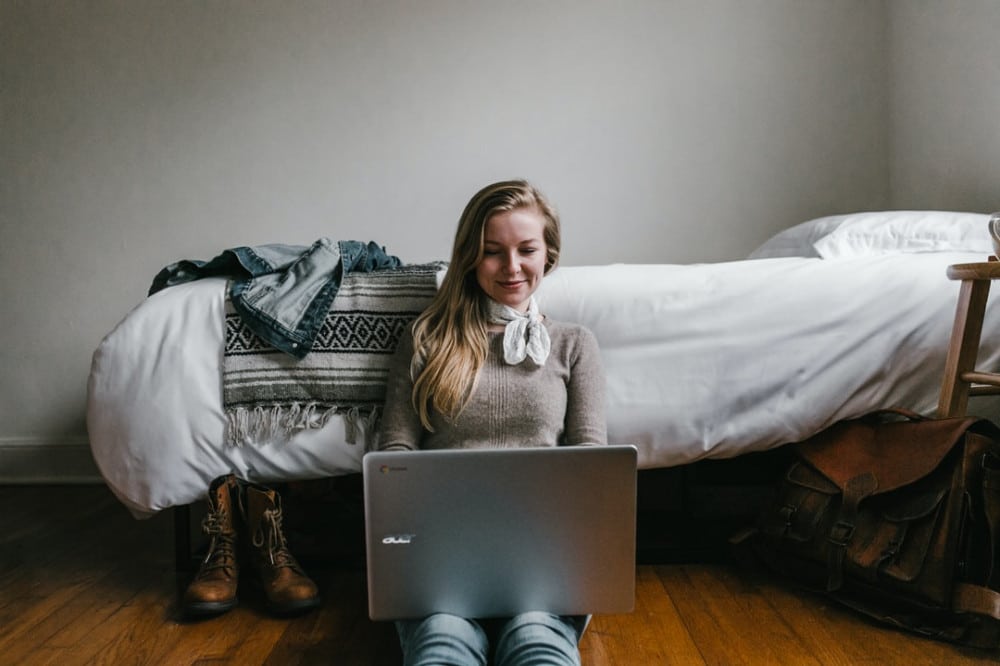 Image of Woman working from the bedroom sitting on the floor with her laptop and shoes off.
